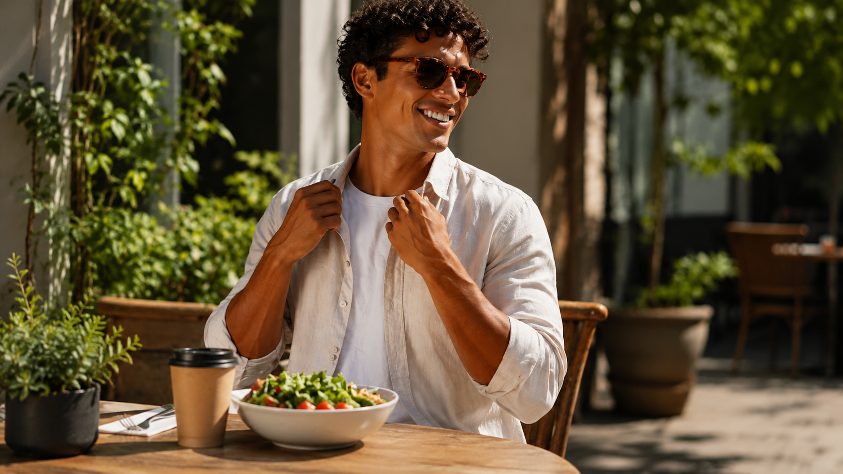 Smiling man wearing sunglasses and a light shirt sits at a sunny outdoor café table with a salad and takeaway coffee, surrounded by lush greenery.